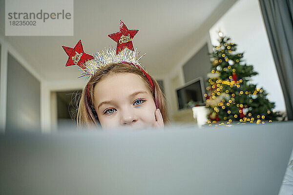 Mädchen mit Weihnachtsstirnband sitzt zu Hause mit Laptop im Schlafzimmer