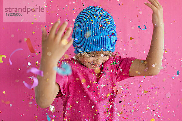 Happy boy wearing blue knit hat playing with confetti against pink background