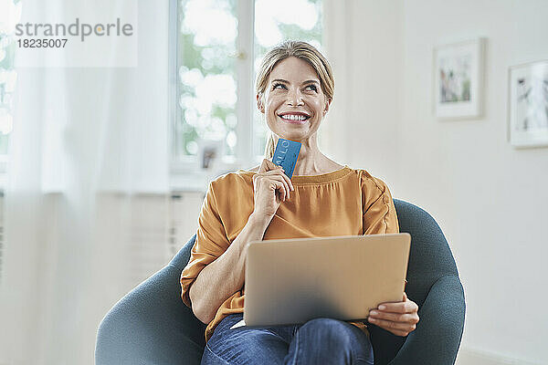 Nachdenklich lächelnde Frau hält zu Hause Kreditkarte und Laptop auf dem Sessel
