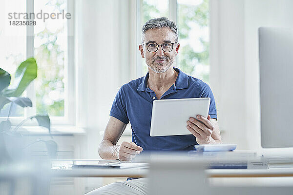 Lächelnder reifer Arzt sitzt mit Tablet-PC am Schreibtisch