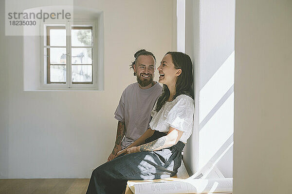 Glücklicher Hipster-Mann mit Freundin sitzt zu Hause auf einem Tisch vor der Wand