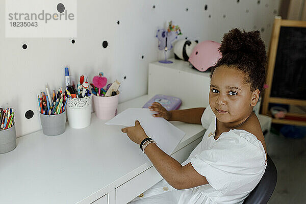 Lächelndes Mädchen mit Papier  das zu Hause am Schreibtisch sitzt