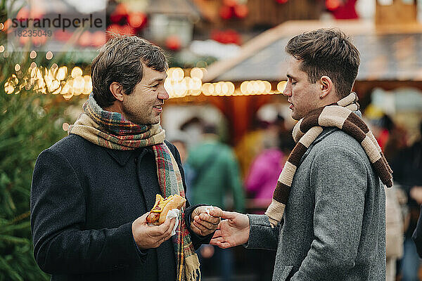 Lächelnder Vater hält Hot Dog in der Hand und unterhält sich mit seinem Sohn  der auf dem Weihnachtsmarkt steht