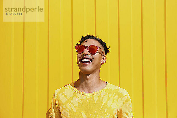 Glücklicher junger Mann mit roter Sonnenbrille vor gelber Wand