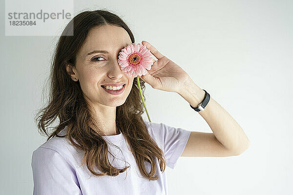 Glückliche Frau bedeckt Auge mit Gerbera-Blume vor weißer Wand