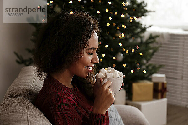 Glückliche Frau hält eine Tasse Marshmallow-Kakao in der Hand und sitzt zu Hause auf dem Sofa
