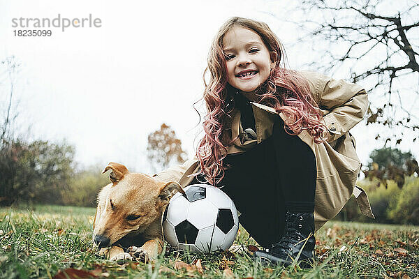 Glückliches Mädchen kauert mit Fußball neben Hund im Park