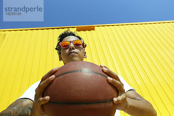 Man wearing red sunglasses holding basketball in front of yellow wall