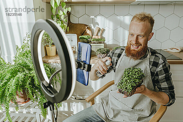 Lächelnder Mann sprüht Wasser auf Microgreens und sitzt vor dem Smartphone