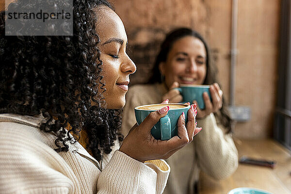 Woman with curly hair smelling coffee by friend in background at cafe
