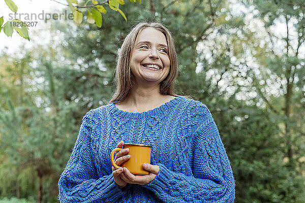Lächelnde Frau im blauen Pullover mit Kaffeetasse und Tagträumen im Freien