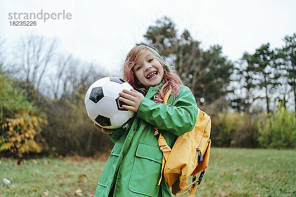 Fröhliches Mädchen steht mit Rucksack und Fußball im Park
