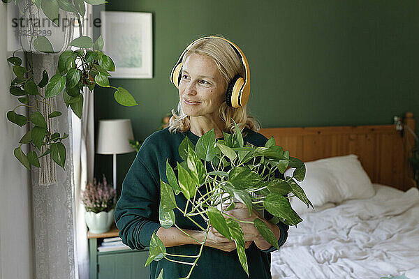 Frau mit Kopfhörern hört Musik und trägt eine grüne Pflanze im Schlafzimmer