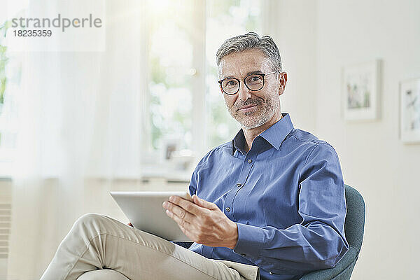 Lächelnder Geschäftsmann hält Tablet-PC im Sessel zu Hause