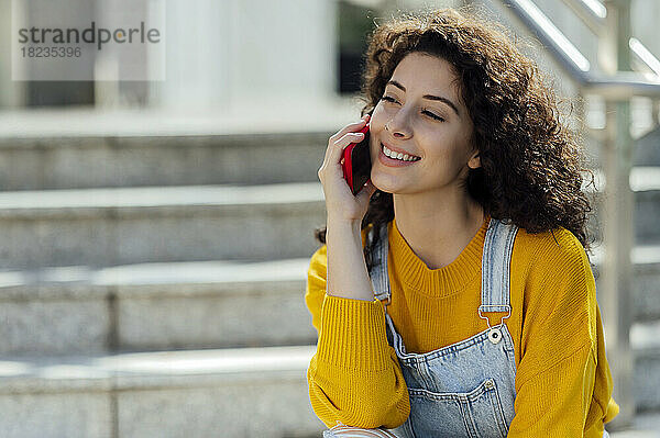 Lächelnde junge Frau mit lockigem Haar  die auf der Treppe über ihr Mobiltelefon spricht