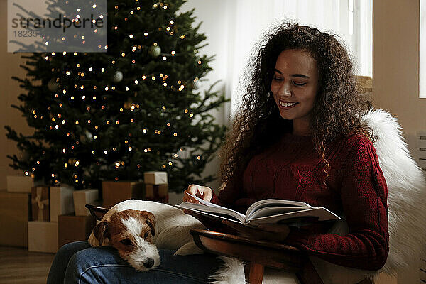 Glückliche Frau sitzt mit Hund und liest zu Hause ein Buch