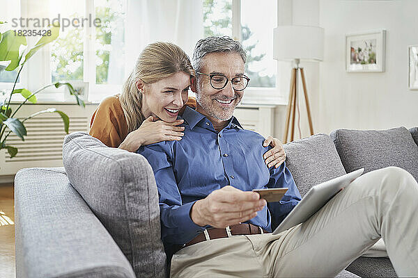Glückliche Frau mit Arm um Mann mit Kreditkarte  Online-Shopping über Tisch-PC auf dem Sofa