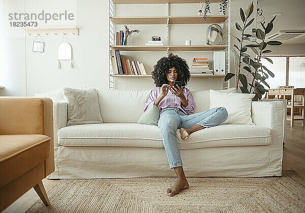 Entspannte Afro-Frau benutzt Smartphone und sitzt zu Hause auf dem Sofa