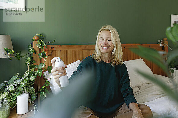 Smiling woman spraying toner on face in bedroom