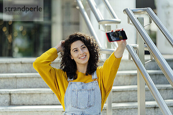 Lächelnde junge Frau mit der Hand im Haar  die auf der Treppe ein Selfie mit dem Mobiltelefon macht