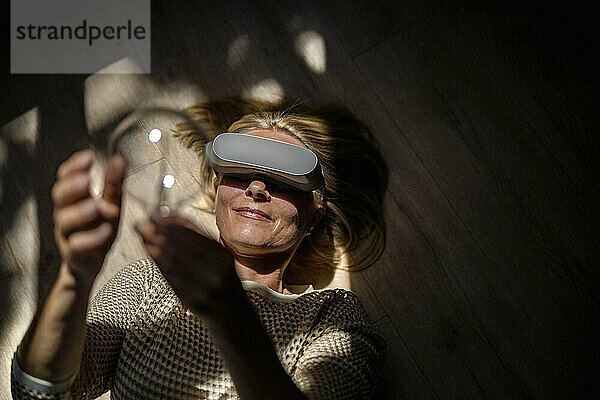 Lächelnde reife Frau mit Glühbirne und Virtual-Reality-Brille zu Hause