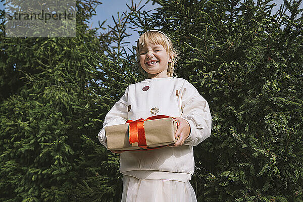 Lächelndes Mädchen mit Geschenkbox  das vor der Tanne steht