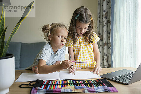 Mädchen zeichnen gemeinsam auf einem Skizzenblock am Laptop am Schreibtisch
