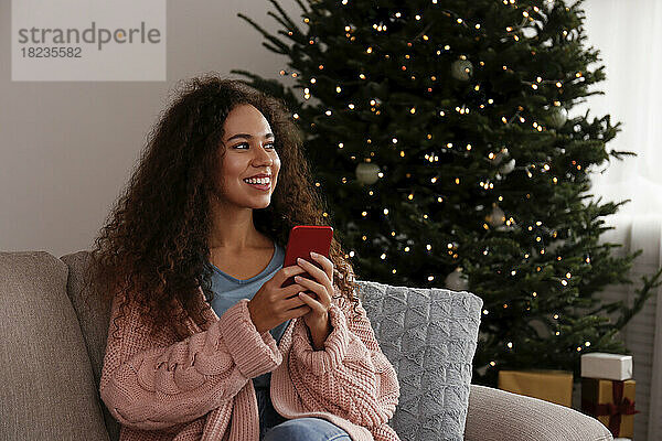 Glückliche junge Frau sitzt mit Smartphone zu Hause