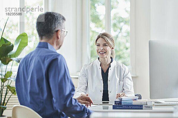 Glückliche Ärztin sitzt am Schreibtisch mit Patientin in der Arztpraxis
