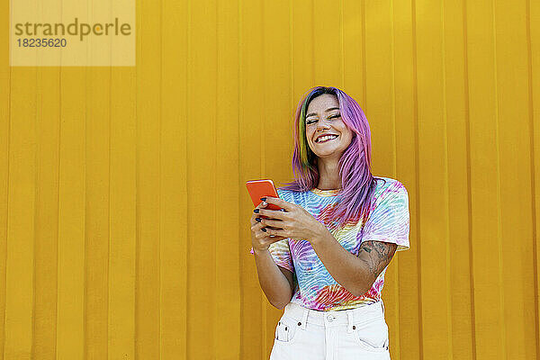 Glückliche Frau hält Mobiltelefon vor gelber Wand