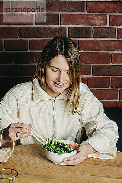 Lächelnde Frau isst Salat mit Stäbchen im Café