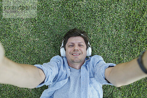 Glücklicher Mann mit Kopfhörern  der ein Selfie macht und im Gras liegt
