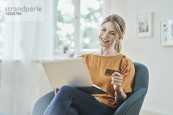 Lächelnde Frau mit Kreditkarte beim Online-Shopping per Laptop im heimischen Sessel