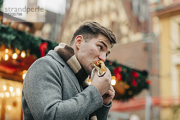 Junger Mann isst Hot Dog und steht auf dem Weihnachtsmarkt
