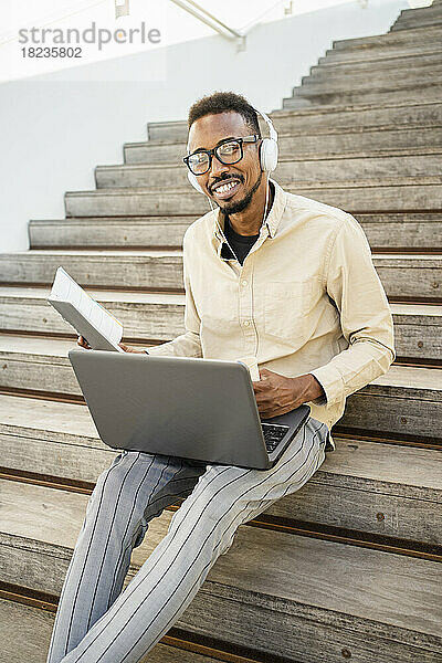 Glücklicher Mann sitzt mit Laptop und trägt Kopfhörer auf Stufen