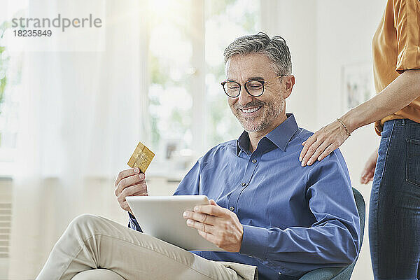 Glücklicher Mann mit Kreditkarte beim Online-Einkauf über Tablet-PC und die Hand einer Frau auf der Schulter zu Hause