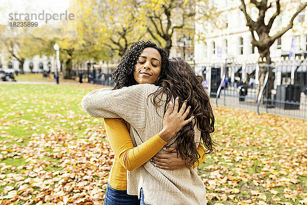Lächelnde Frau mit geschlossenen Augen umarmt Freundin im Herbstpark