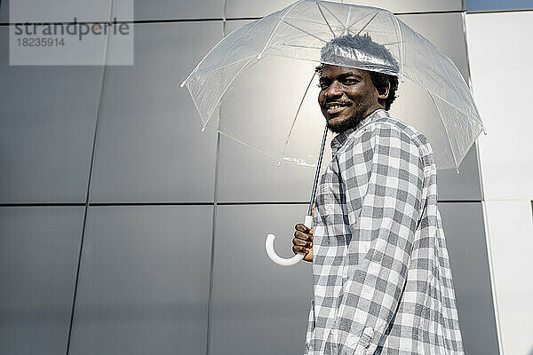 Lächelnder Mann hält durchsichtigen Regenschirm vor der Wand