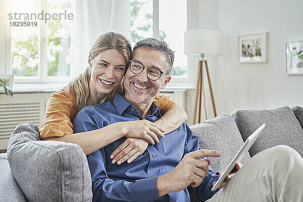 Glückliche Frau umarmt Mann mit Tablet-PC von hinten auf dem heimischen Sofa