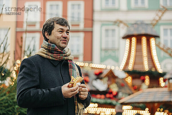 Lächelnder reifer Mann steht mit Hot Dog auf dem Weihnachtsmarkt