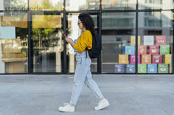 Junge Frau mit Rucksack benutzt Smartphone neben Glasgebäude auf Fußweg