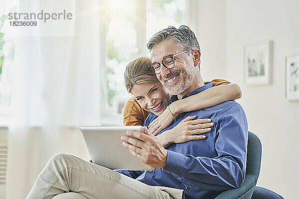 Glücklicher Mann zeigt Frau im Sessel zu Hause Tablet-PC
