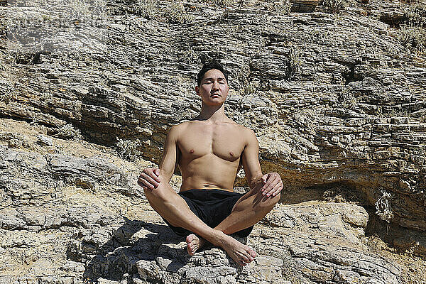 Nackter Oberkörper junger Mann mit geschlossenen Augen meditiert an einem sonnigen Tag auf einem Felsen