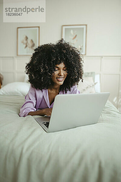Lächelnde Frau mit Laptop auf dem Bett im Schlafzimmer liegend