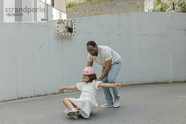 Vater schiebt Tochter  die auf Skateboard vor Wand sitzt