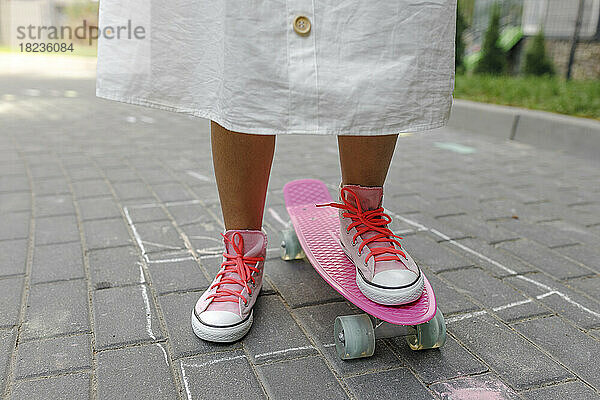 Mädchen mit rosa Skateboard steht auf Fußweg