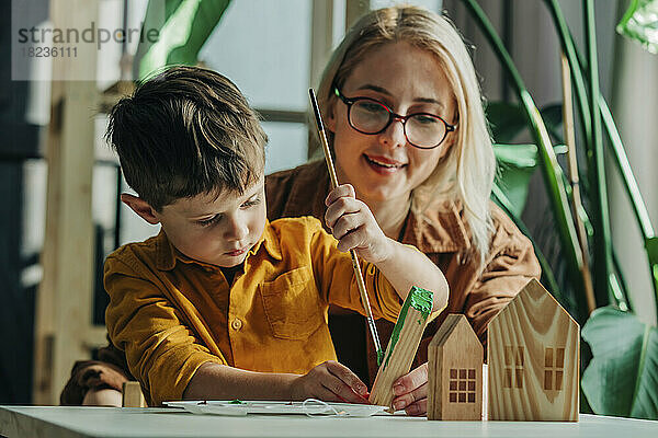 Lächelnde Mutter und Sohn bemalen Holzmodellhäuser am Tisch