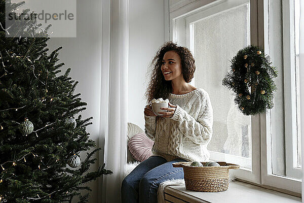 Glückliche Frau  die zu Hause auf der Fensterbank mit einer Tasse Marshmallow-Kakao sitzt