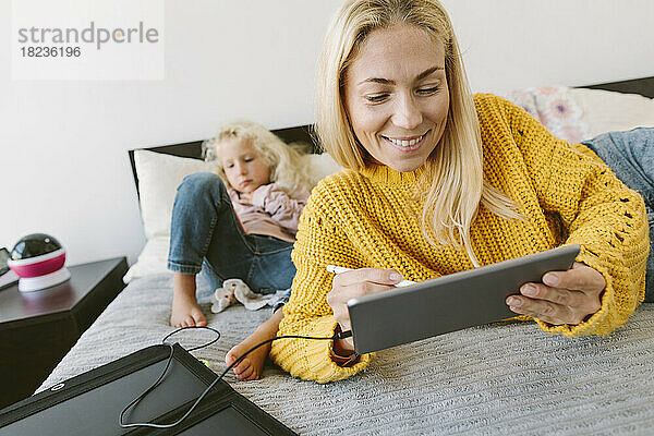 Glückliche Frau mit Tablet-PC  die sich auf das Bett stützt  mit ihrer Tochter im Hintergrund