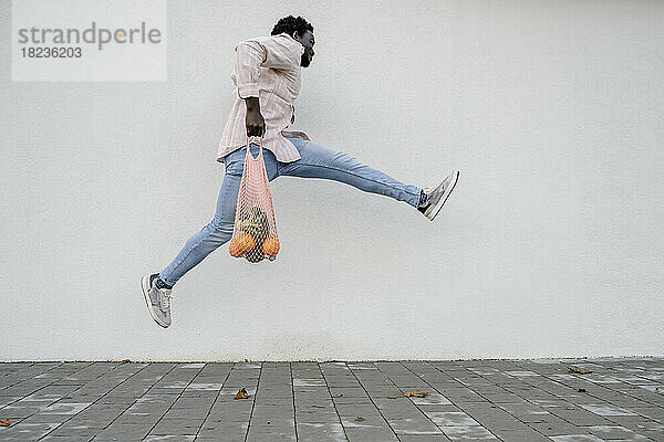 Junger Mann mit Netzbeutel springt vor weiße Wand